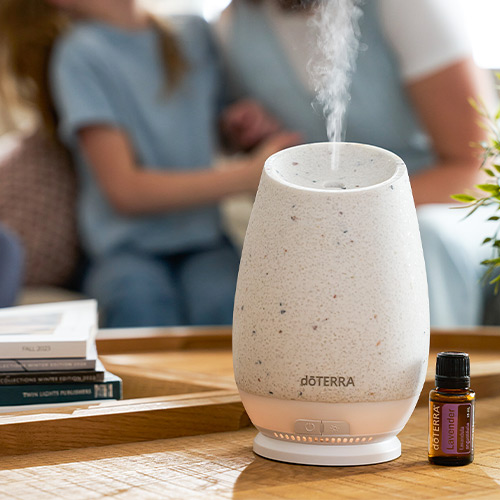 A close-up of a doTERRA Roam Diffuser and Lavender Essential Oil on a wooden table, and blurred people in the background.
