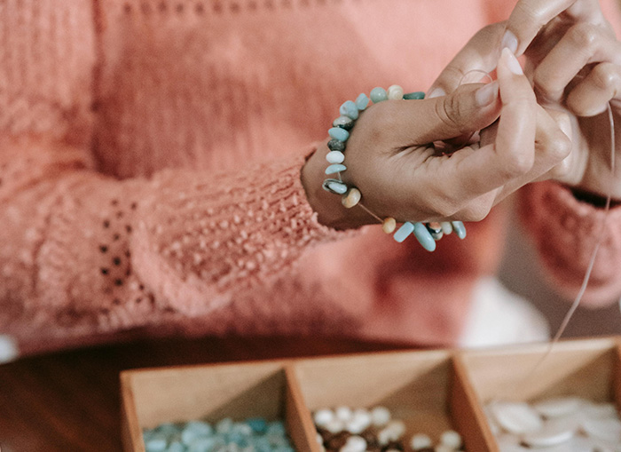 A woman sitting at a table holding a half-beaded turquoise bracelet around her wrist as she adds more bead. A tray of beads sits below her hands. 