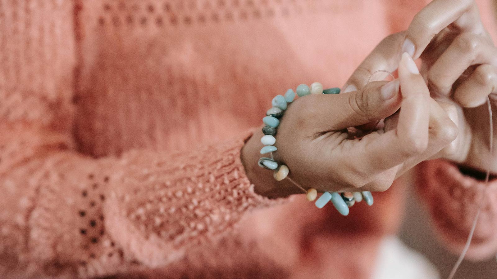 A woman sitting at a table holding a half-beaded turquoise bracelet around her wrist as she adds more bead. A tray of beads sits below her hands. 