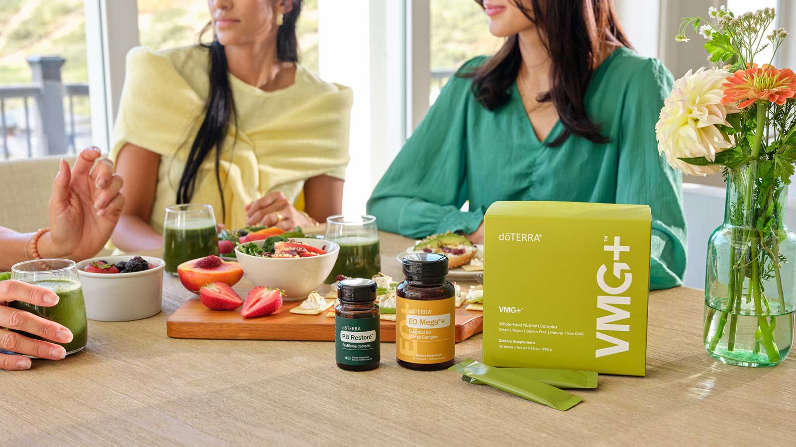 Three women sitting at a table with fruit, VMG+, PB Restore, and EO Mega+ scattered on the table. 