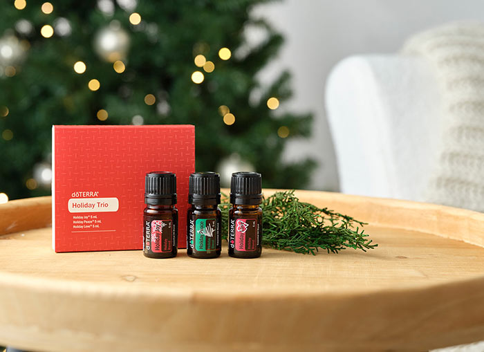 Holiday Trio on round, wooden table. A white couch with a cream blanket draped over it and a lit Christmas tree in the background.