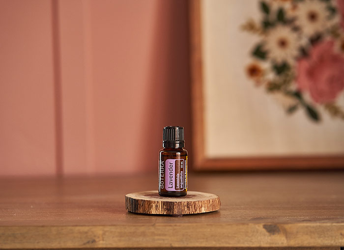 doTERRA Lavender essential oil seated on thin bark platform on a wooden table. A pink wall and floral needlework framed in the background. 