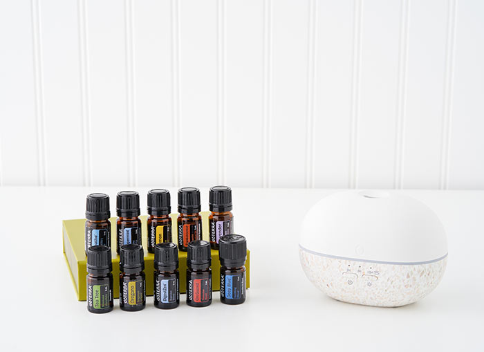 On a white background, each of the doTERRA Healthy Start Kit essential oils is organized in two lines, the first row on top of a green essential oil storage box. The doTERRA Pebble Diffuser sitting to the side.