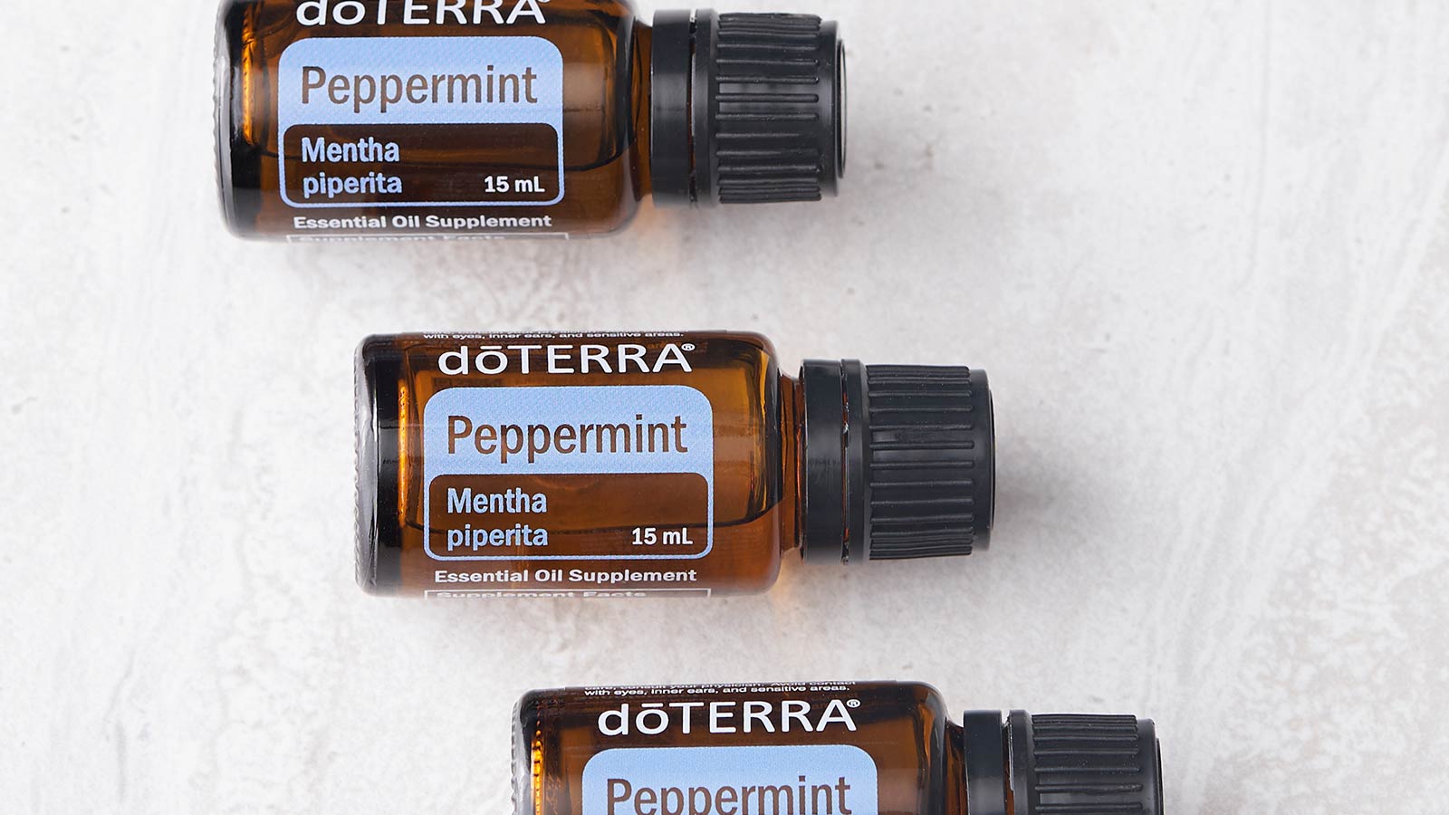 Three doTERRA Peppermint essential oils laying in a vertical, off-centered line on a white surface. 