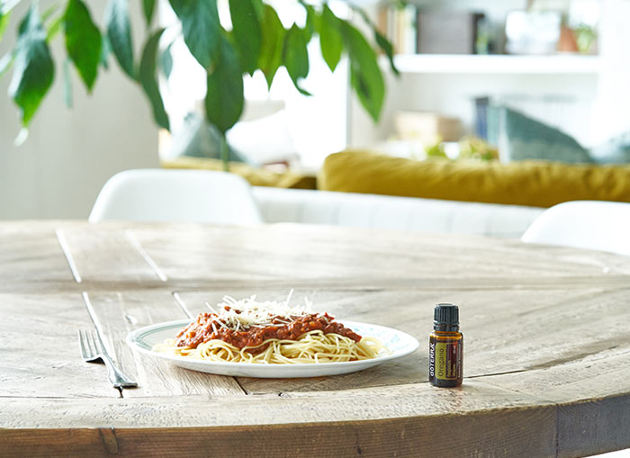 doTERRA Oregano essential oil sitting on wooden, round, kitchen table, beside a plate of spaghetti. A large, green plant, couch, and living room in the background. 