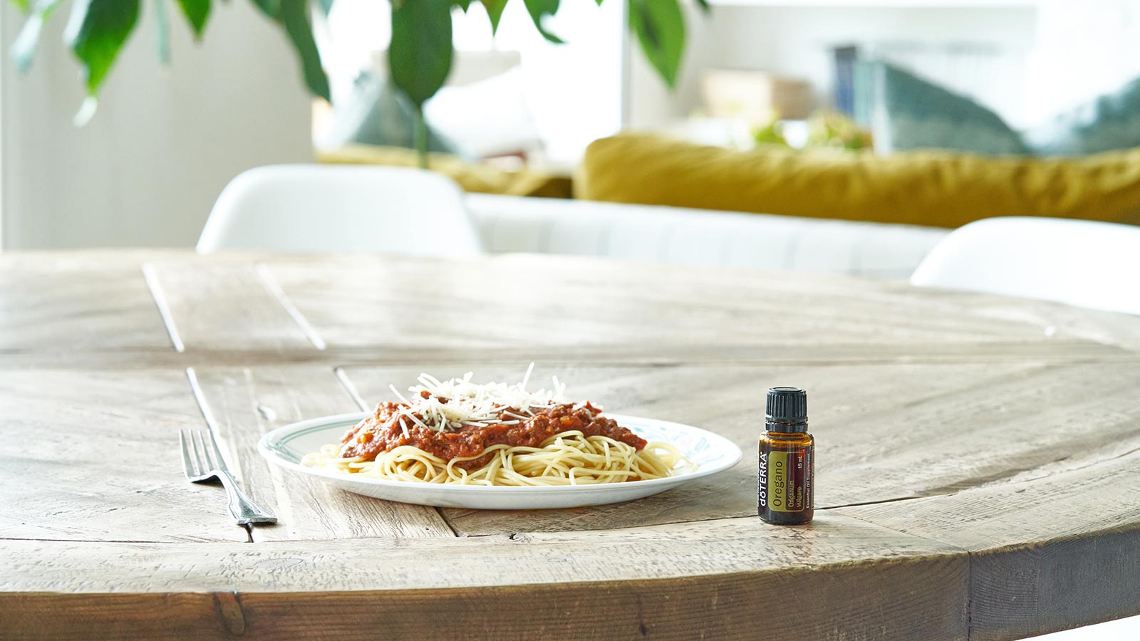 doTERRA Oregano essential oil sitting on wooden, round, kitchen table, beside a plate of spaghetti. A large, green plant, couch, and living room in the background. 