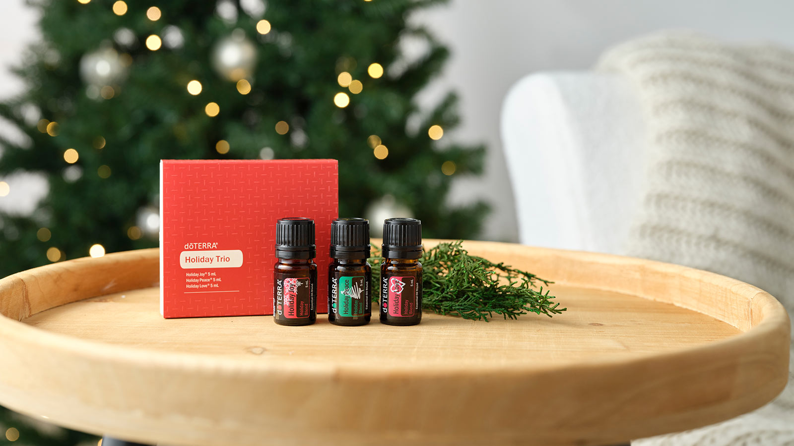Holiday Trio on round, wooden table. A white couch with a cream blanket draped over it and a lit Christmas tree in the background.