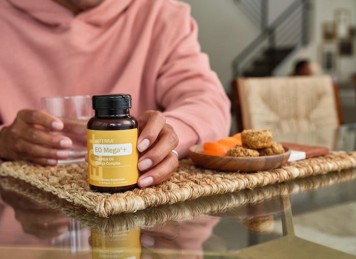 Woman’s hand around EO Mega+ Bottle and her other hand around a glass water, lying on a woven placement. Small plate of food, sitting to the side. 