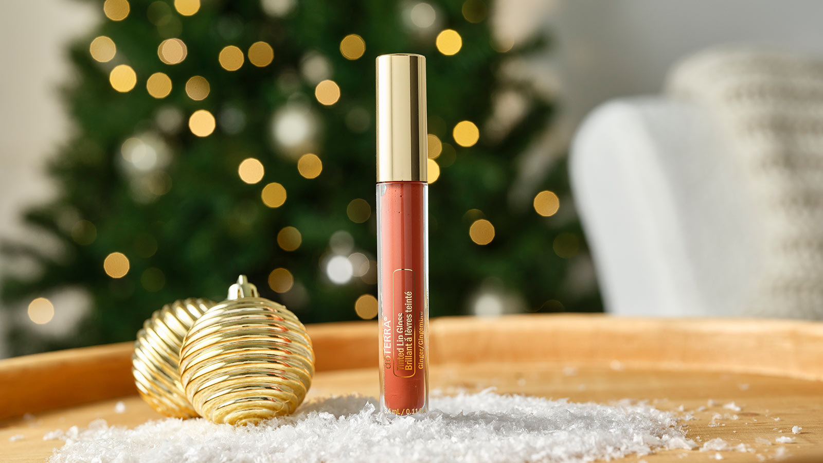 Tinted Lip Gloss standing on a round, wooden table with two gold ornaments and fake snow surrounding it. A lit Christmas tree and chair are faded in the background.