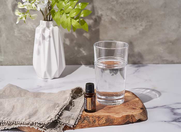 doTERRA Ginger essential oil sitting on wooden board with a cup of water and towel beside it. A bouquet of white flowers and greenery are in the background. 