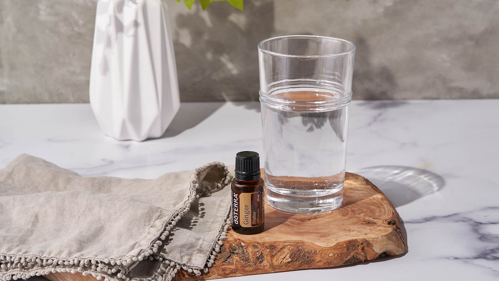 doTERRA Ginger essential oil sitting on wooden board with a cup of water and towel beside it. A bouquet of white flowers and greenery are in the background. 