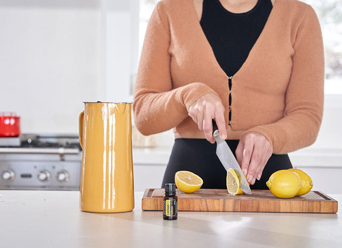 A woman cutting lemons on a wooden cutting board, with a pitcher and doTERRA Lemon essential oil sitting beside the cutting board on the counter. 