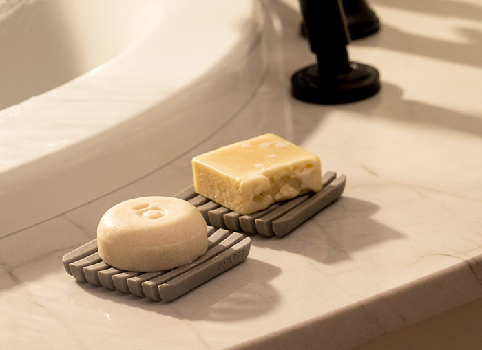 doTERRA Shampoo and Conditioner Strengthening Bars sitting on gray soap dishes, placed on a marble counter near a bathtub. 