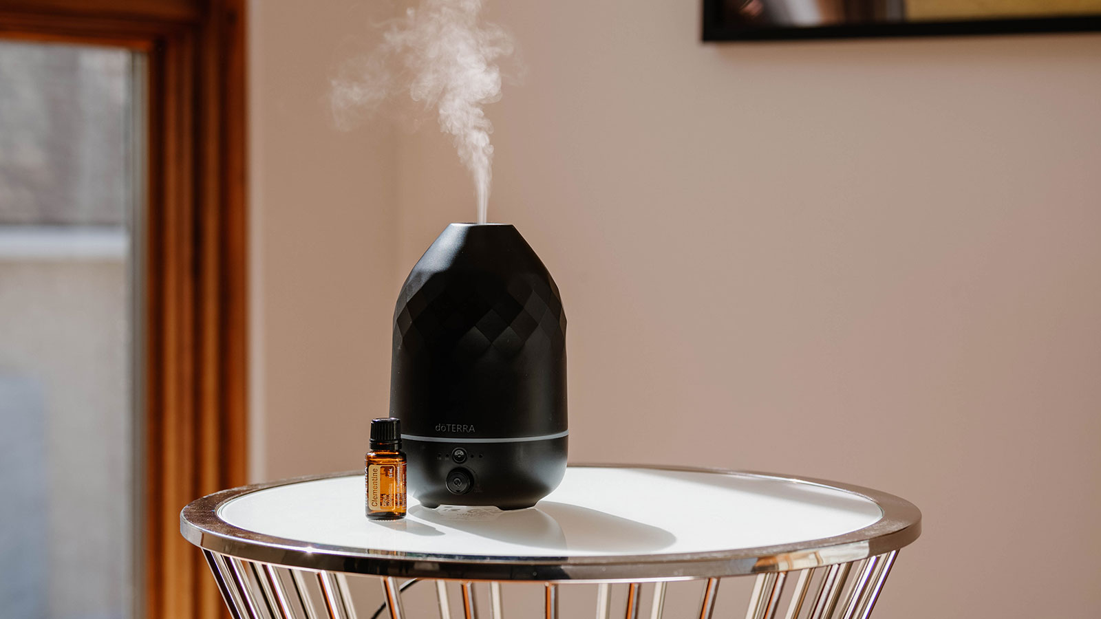 Volo Onyx Diffuser sitting on a white, round table. Mist coming out. A doTERRA Clementine essential oil bottle sitting beside it. 