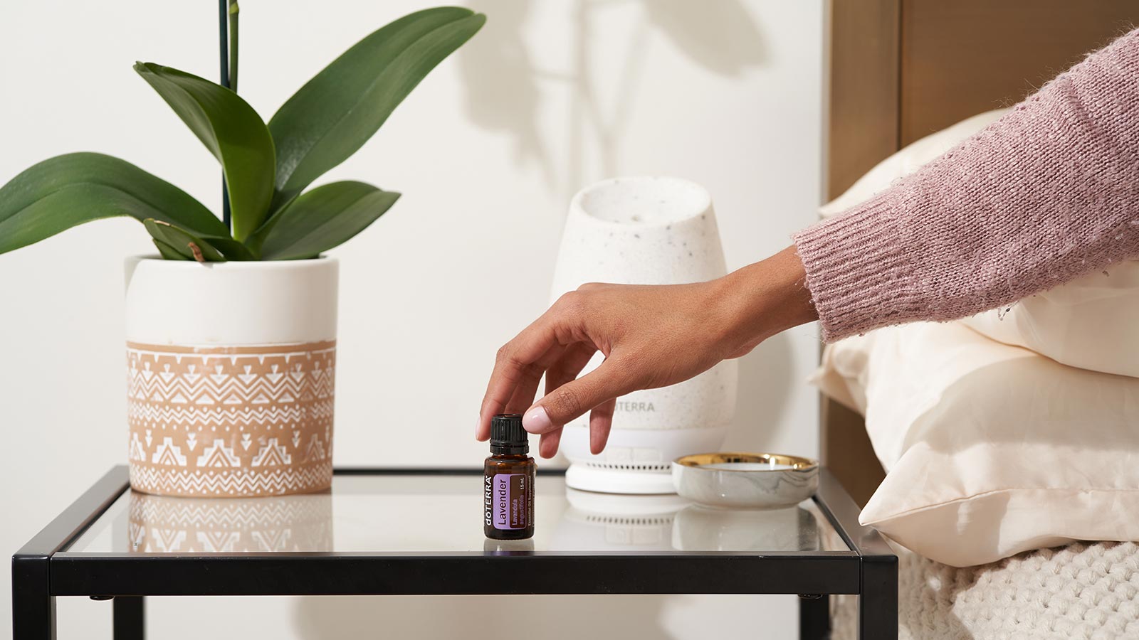 Woman, sitting on a bed, reaching for doTERRA Lavender essential oil on nightstand table. The doTERRA Roam Diffuser and a large green plant behind the oil.  