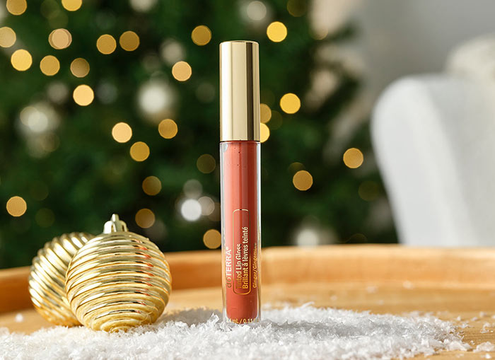 Tinted Lip Gloss standing on a round, wooden table with two gold ornaments and fake snow surrounding it. A lit Christmas tree and chair are faded in the background.