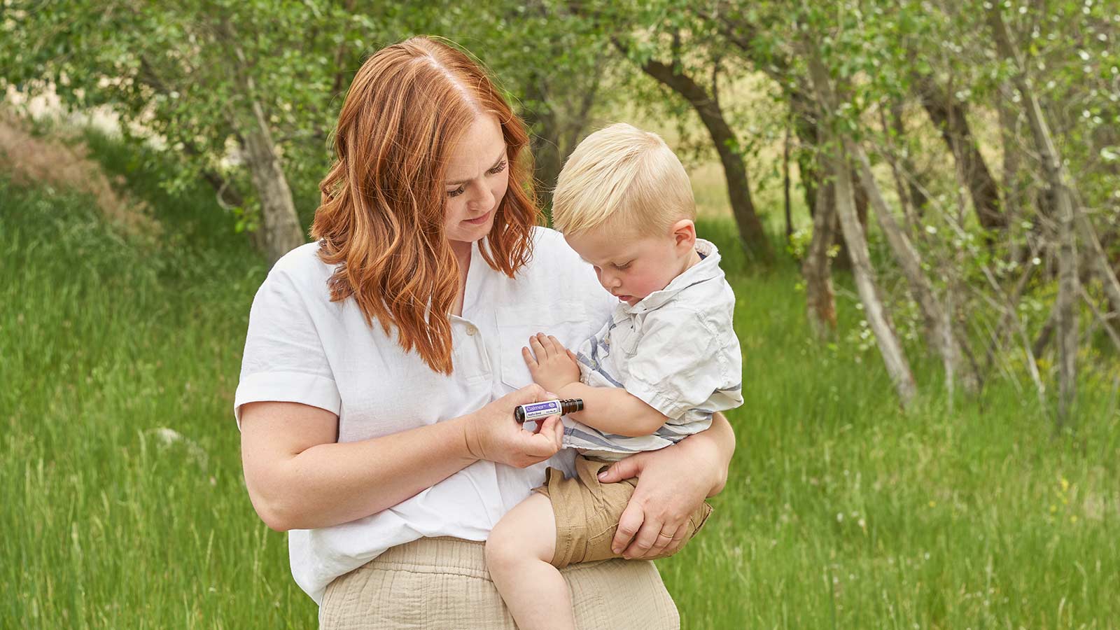 A woman stands outside in green grass, holding her son, while applying doTERRA Calmer Touch to her son’s arm. 