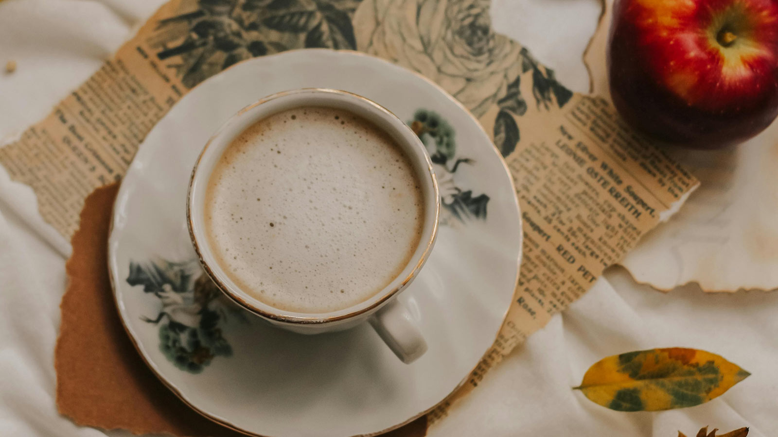 An overhead shot of a cup of tea sitting on a saucer. The saucer is placed on a ripped newspaper and a few leaves sit beside it. A large, red apple sits off to the side. 