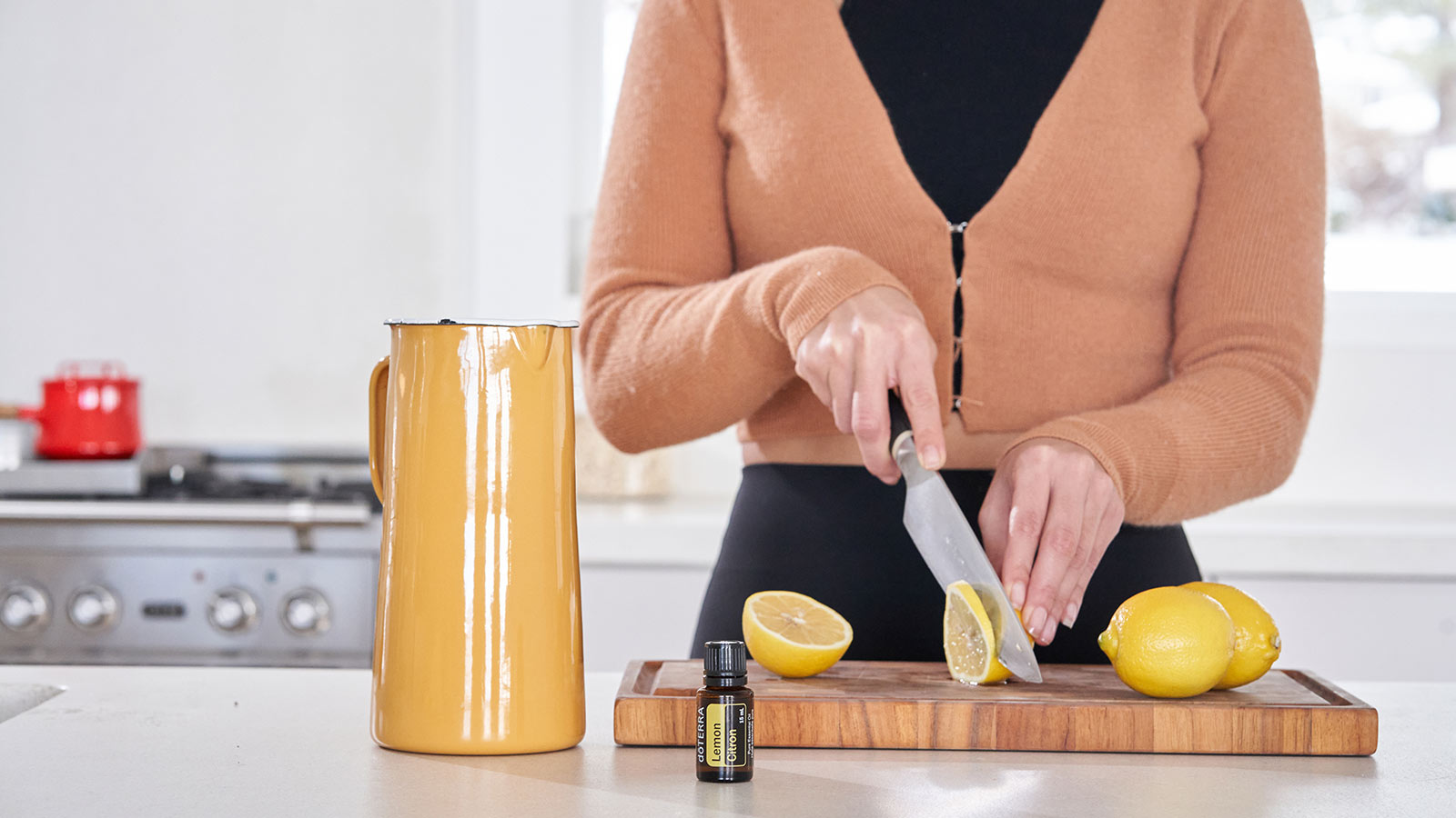 A woman cutting lemons on a wooden cutting board, with a pitcher and doTERRA Lemon essential oil sitting beside the cutting board on the counter. 