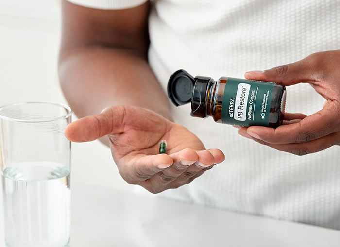 Man holding the PB Restore ProBiome Complex bottle, gently pouring the pills out. A clear glass of water sits on a white counter. 