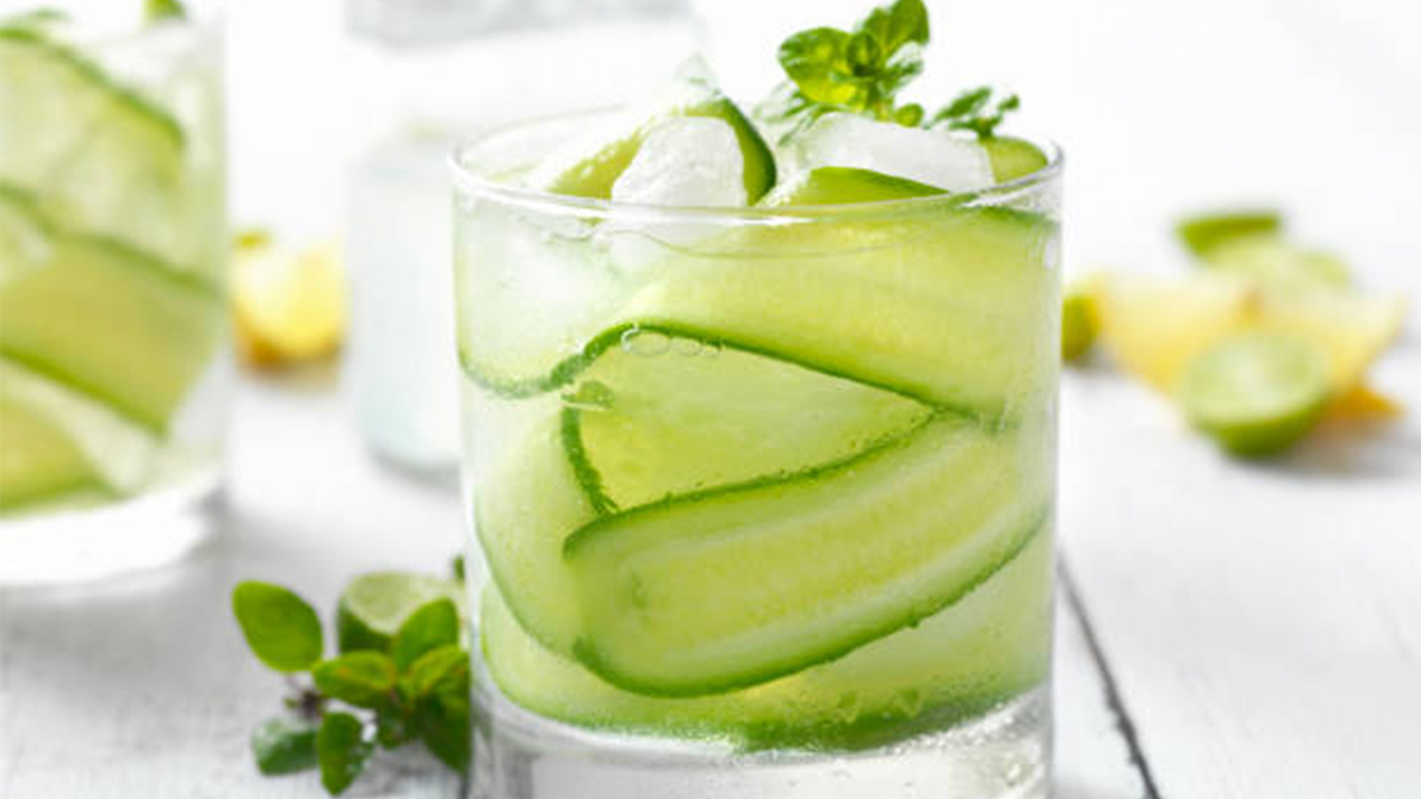 A glass of thinly sliced cucumber, mint leaves, and sparkling water sits on a gray surface. An additional cup and lemon and lime fruit slices sit in the background. 