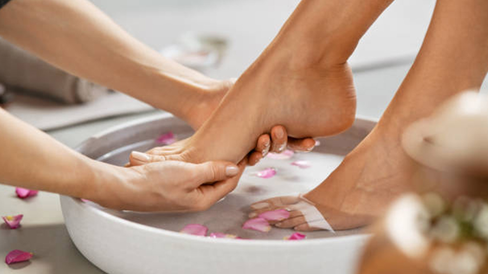 Someone has their feet gently placed in a round-white bowl and peddle-covered water. One of their feet is being lifted by someone else’s hand. The scenery showcases a spa-like experience. 