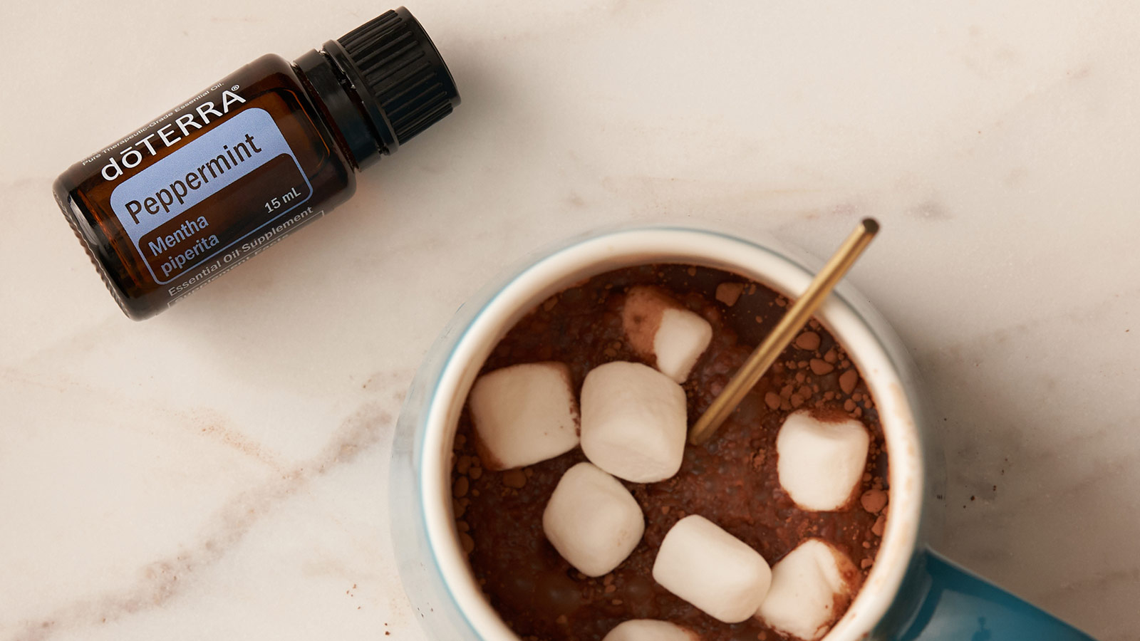 doTERRA Peppermint essential oil lying flat on a countertop. A mug, filled with hot chocolate, marshmallows, and a spoon, sit beside it. 