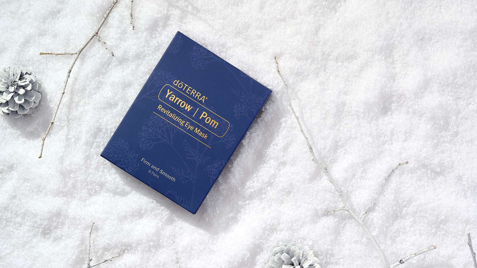 Yarrow Pom Eye Mask laying on a soft, snow-like surface surrounded by white twigs and pinecones.