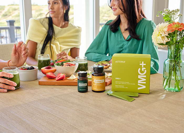 Three women sitting at a table with fruit, VMG+, PB Restore, and EO Mega+ scattered on the table. 