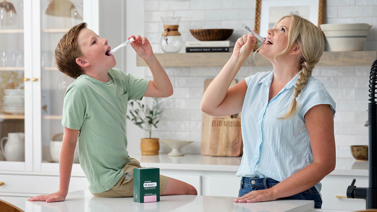 Son and mother are pouring PB Assist+ ProBiome Gut Complex sachets into their mouths in a primarily white kitchen. The son sits on the counter, with the mother standing beside him.