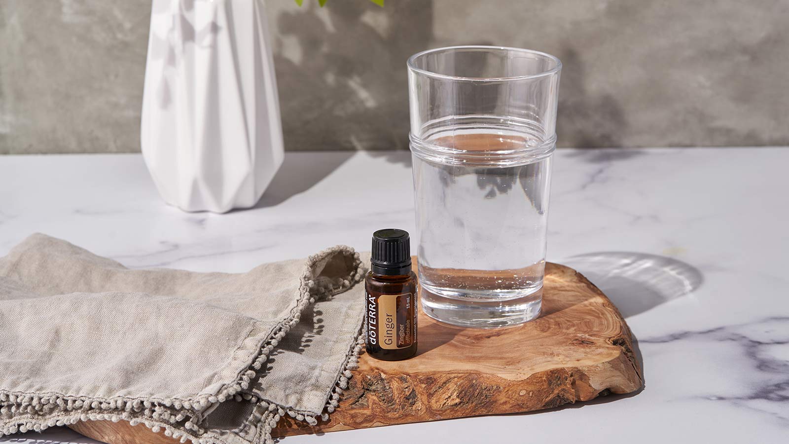 doTERRA Ginger essential oil sitting on a wooden board, with a gray towel and clear glass of water on either side. A vase of flowers sits in the background.