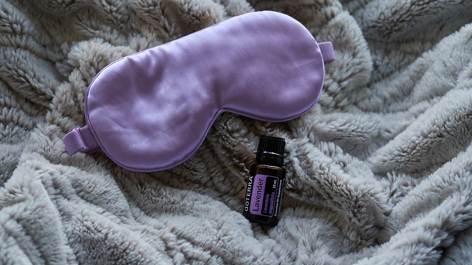doTERRA Lavender essential oil lays on a fluffy, gray blanket, with a purple sleep mask above it.  