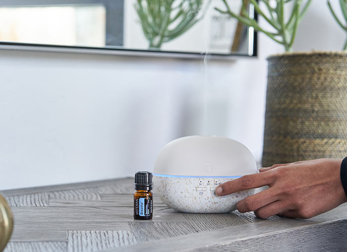 A woman pressing her finger against the doTERRA Pebble Diffuser as mist comes out from the top. Breathe essential oil blend sits to the side of the diffuser. 
