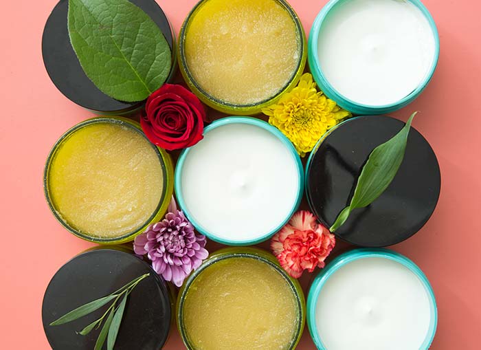 Overhead shot of nine bottles of lotion and body scrub with some lids open and some closed. The containers create a square with leaves and flowers between them.  