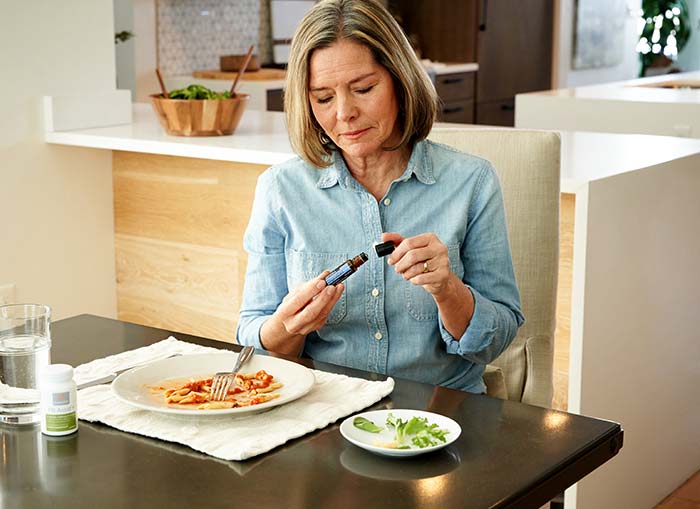 A woman sitting at a kitchen table, finishing up her meal. She is using both hands to unscrew doTERRA DigestZen Touch. PB Assist sits near a glass of water on the black tabletop. 