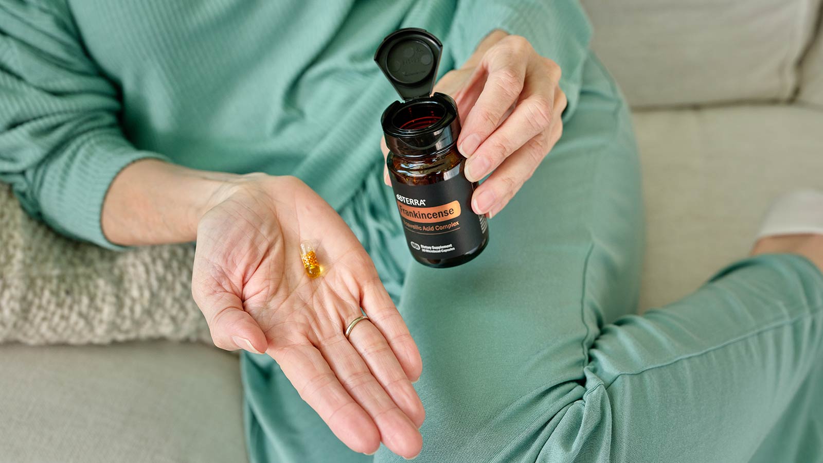 A person laying on a couch holding the Boswellic Acid Complex bottle in one hand and holding a capsule in the other.