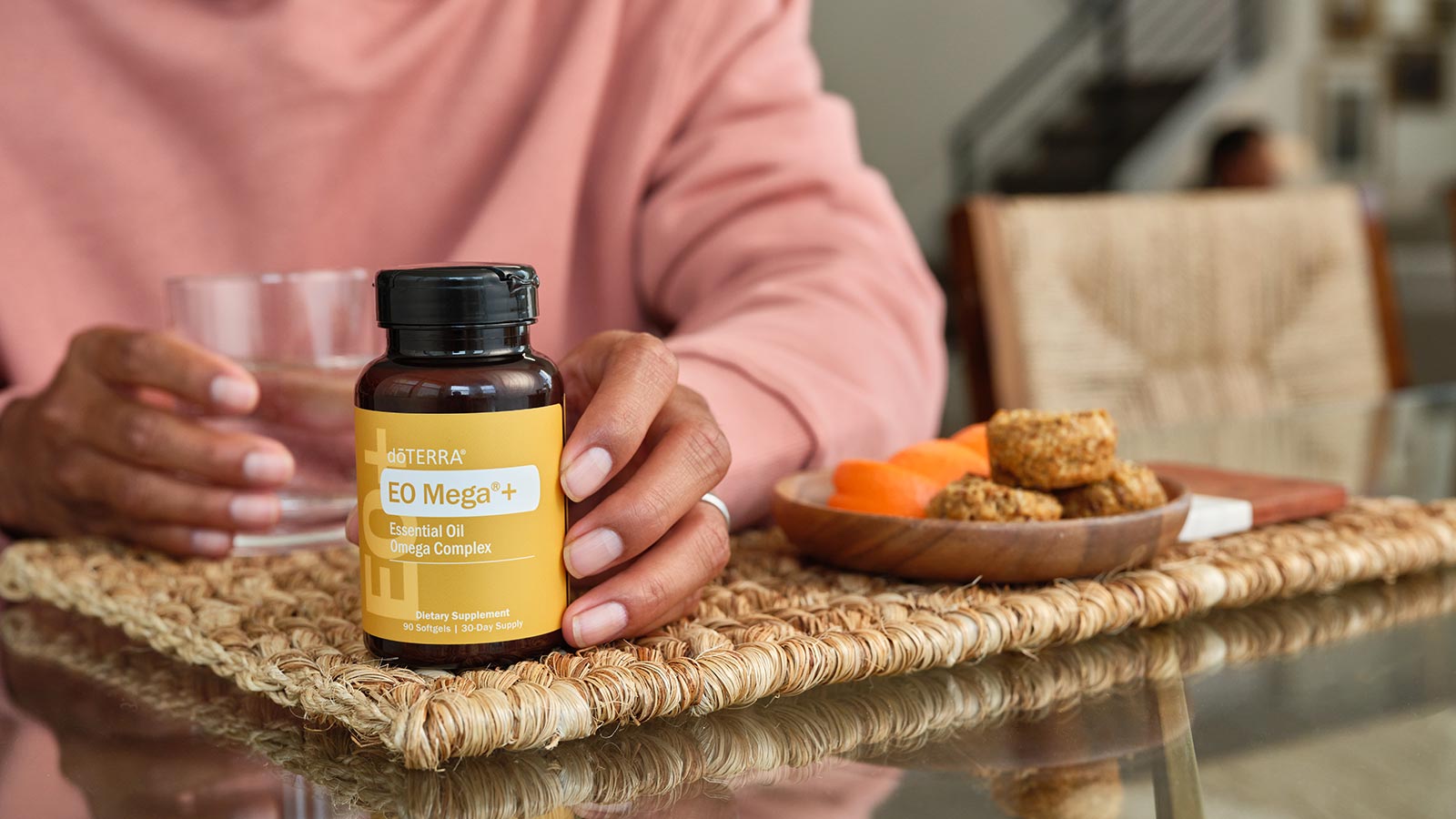 Woman’s hand around EO Mega+ Bottle and her other hand around a glass water, lying on a woven placement. Small plate of food, sitting to the side. 