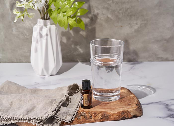 doTERRA Ginger essential oil sitting on a wooden board, with a gray towel and clear glass of water on either side. A vase of flowers sits in the background.