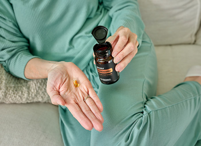 A person laying on a couch holding the Boswellic Acid Complex bottle in one hand and holding a capsule in the other.
