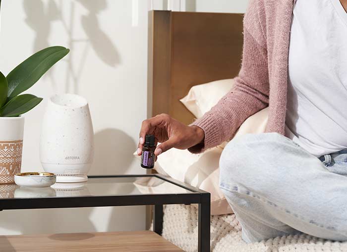 A woman sitting on a bed, a nightstand beside her with the doTERRA Roam Diffuser on the nightstand. She is lifting Lavender essential oil from the table. 