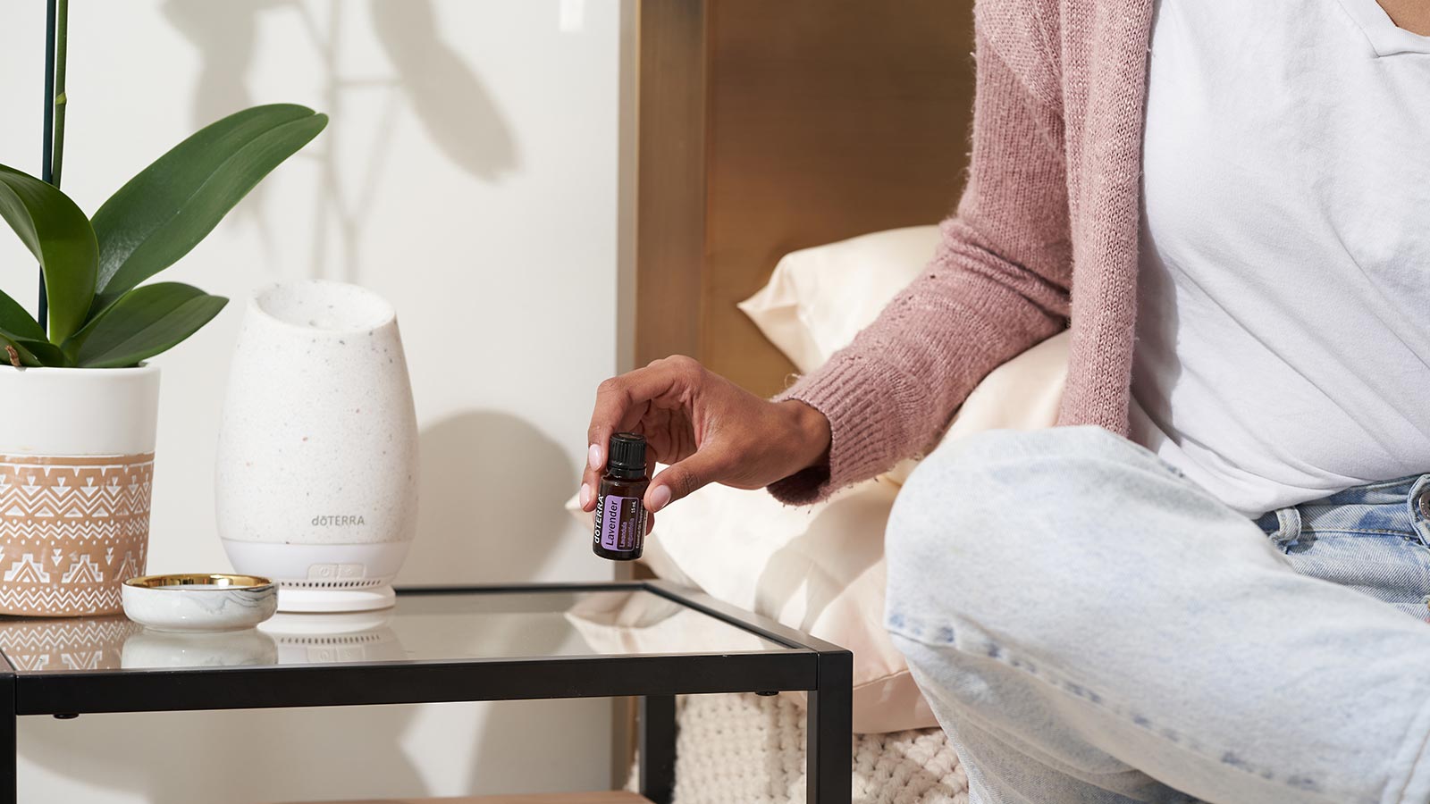 A woman sitting on a bed, a nightstand beside her with the doTERRA Roam Diffuser on the nightstand. She is lifting Lavender essential oil from the table. 