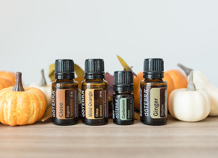 doTERRA essential oils of Clove, Wild Orange, Cardamom, and Ginger all lined up on a brown table. Small orange and white pumpkins are arranged behind them. 