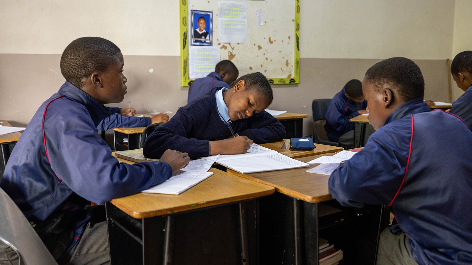 Several children in navy blue school uniforms sitting at desk clumps busy with their schoolwork.