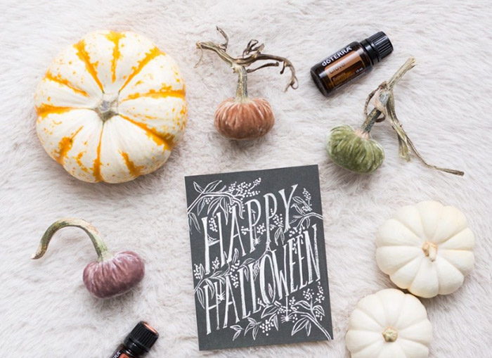 A black Happy Halloween card placed on a white, soft surface, surrounded by pumpkins and essential oil bottles. 