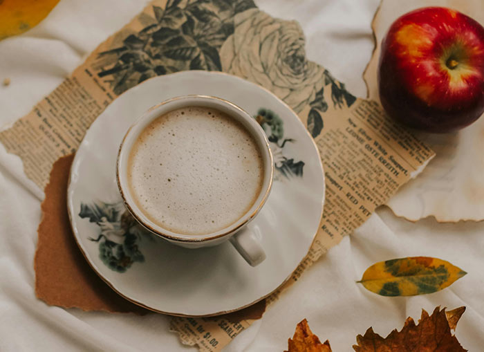 An overhead shot of a cup of tea sitting on a saucer. The saucer is placed on a ripped newspaper and a few leaves sit beside it. A large, red apple sits off to the side. 