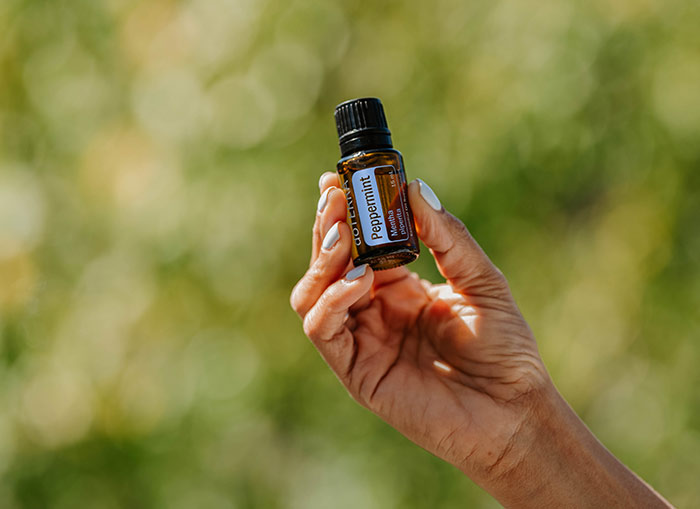 Someone with blue painted fingernails, holding up a bottle of doTERRA Peppermint essential oil against a green faded background. 