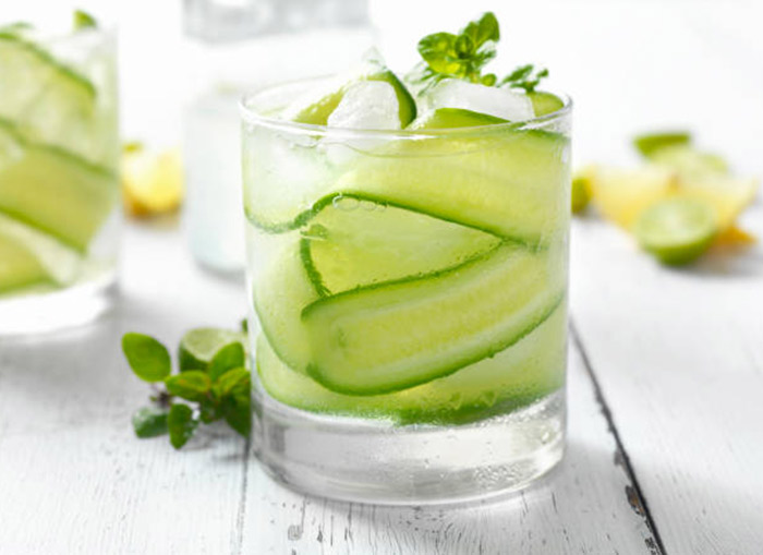 A glass of thinly sliced cucumber, mint leaves, and sparkling water sits on a gray surface. An additional cup and lemon and lime fruit slices sit in the background. 