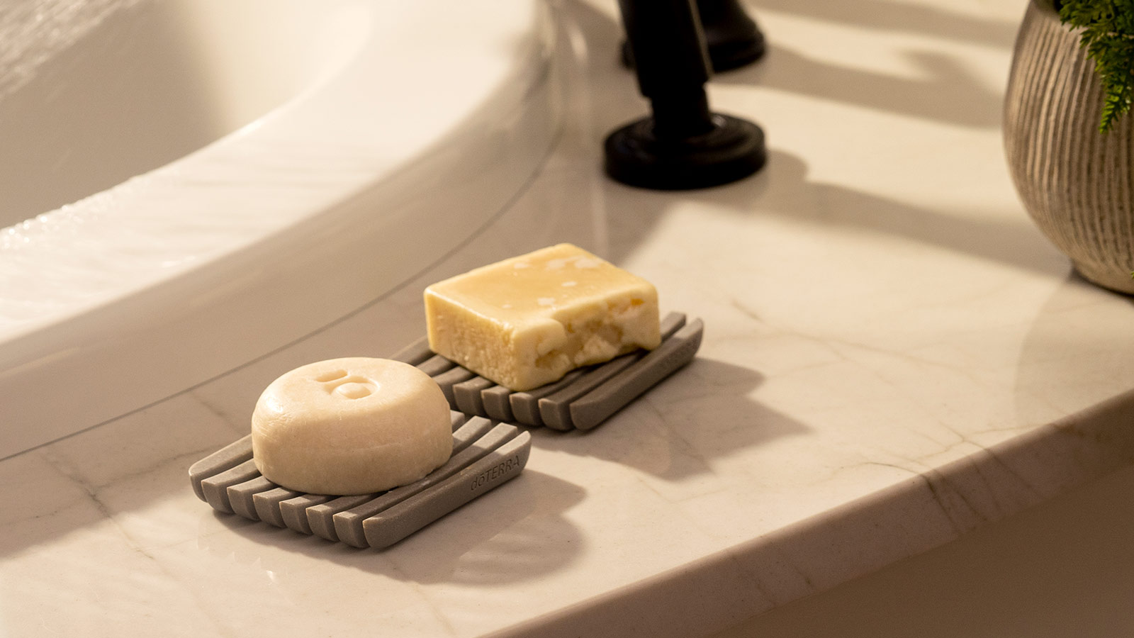 doTERRA Shampoo and Conditioner Strengthening Bars sitting on gray soap dishes, placed on a marble counter near a bathtub. 