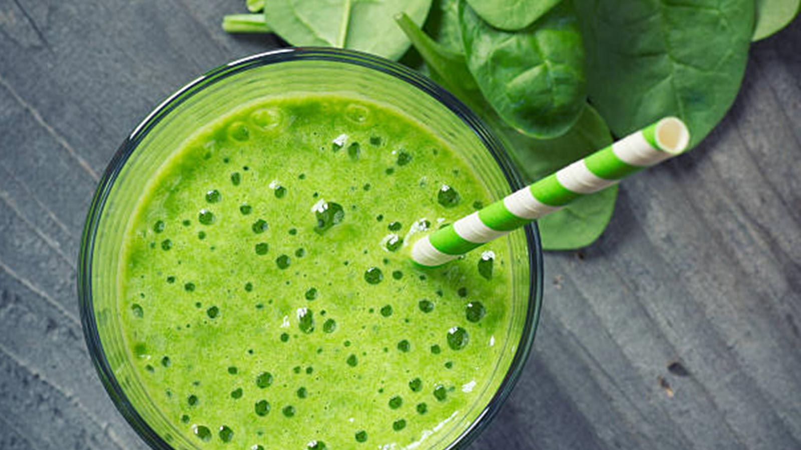 An overhead shot of a green drink in a glass cup with a green and white striped straw in the cup. The cup is on a gray surface with spinach sitting to the side. 