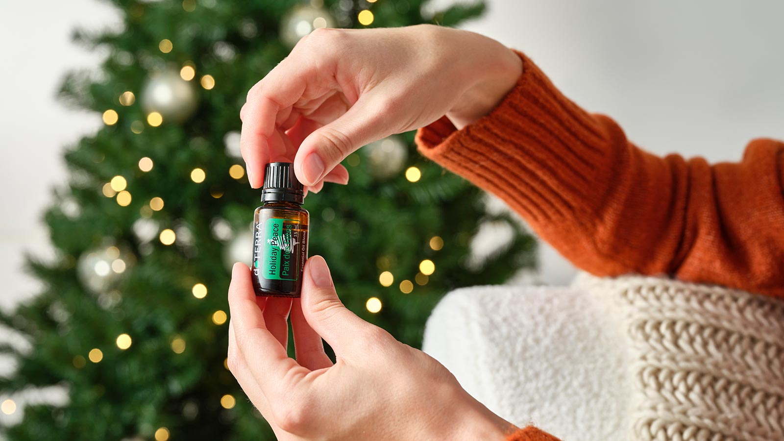 Woman holding doTERRA Holiday Peace essential oil blend, while resting her elbow on the armrest of a white couch. A lit Christmas tree in the background. 